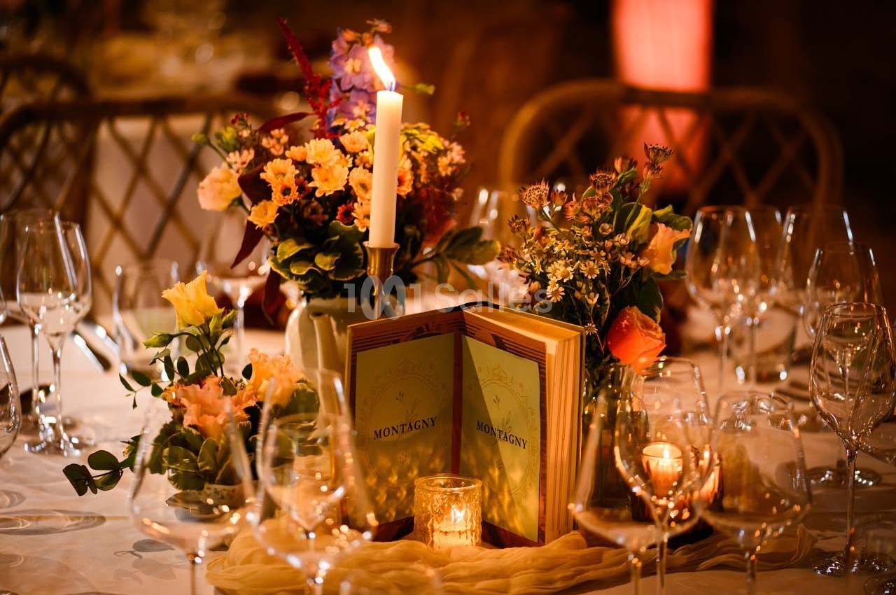 Table élégamment dressée avec bougies, fleurs colorées et menus ’Montagny’ entourés de verres à vin.