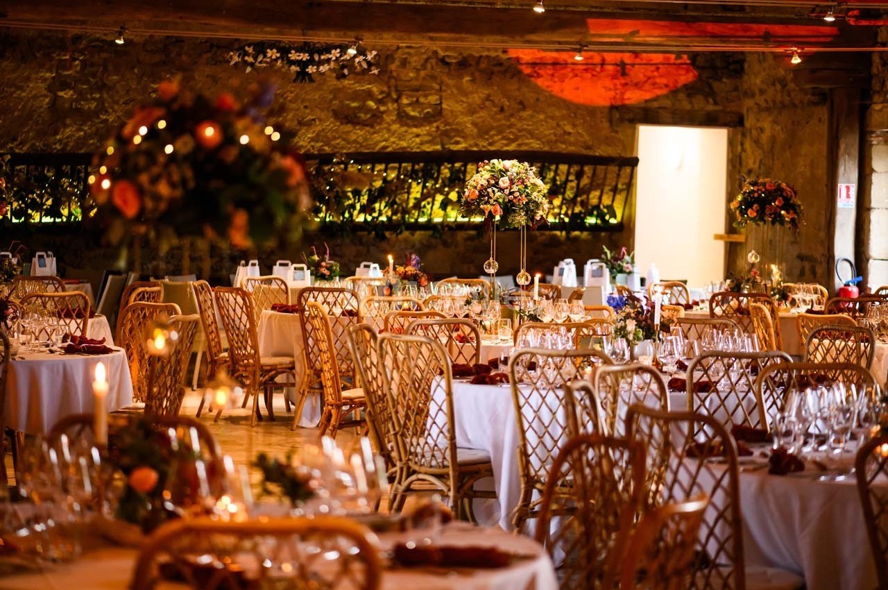 Salle de réception décorée avec des tables rondes, nappes blanches, chaises en rotin et arrangements floraux colorés.