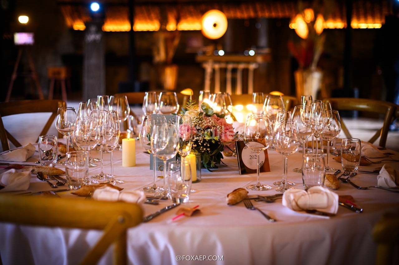 Table élégamment dressée avec nappes blanches, verres à vin, bougies et centre de table floral dans une ambiance chaleureuse.