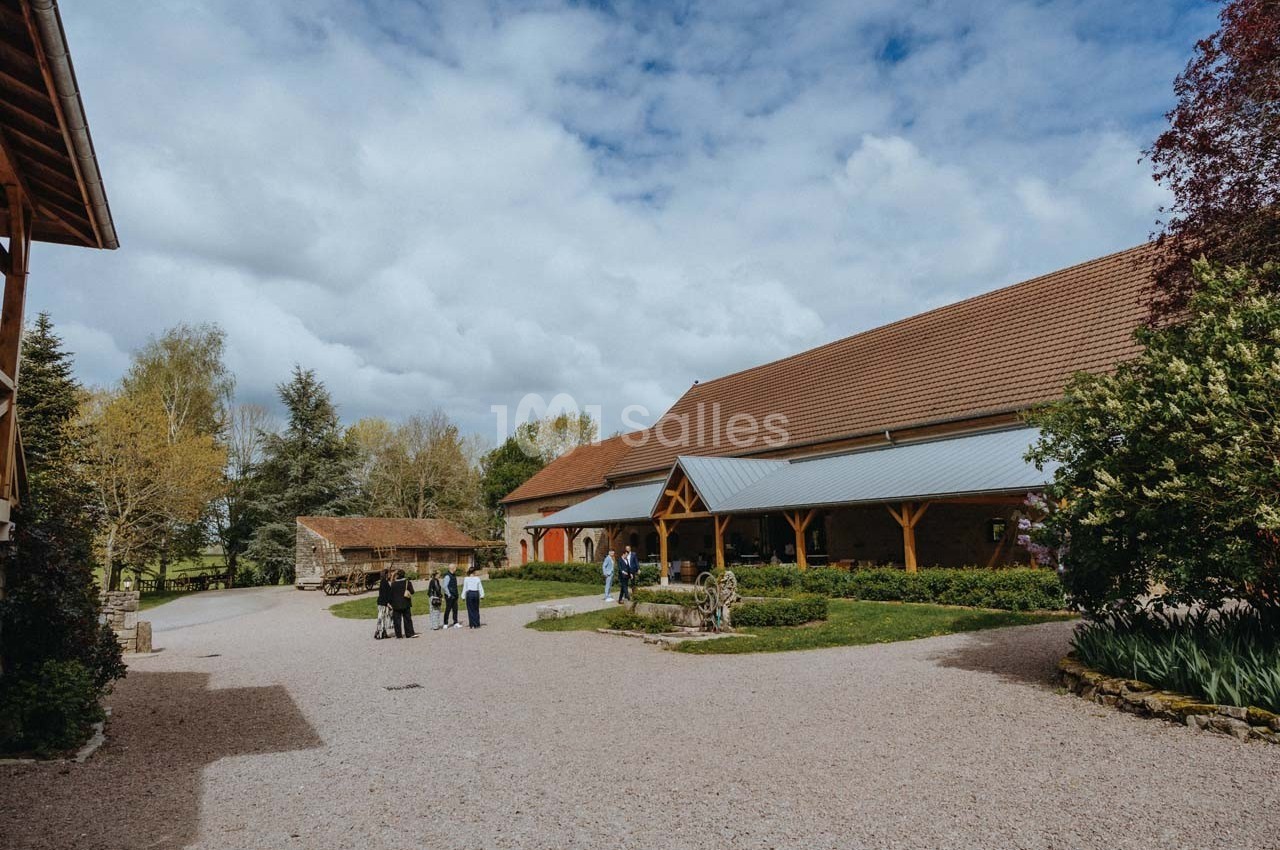 Cour d'une ferme avec bâtiments en pierre et en bois, quelques arbres, et un petit groupe de personnes discutant.