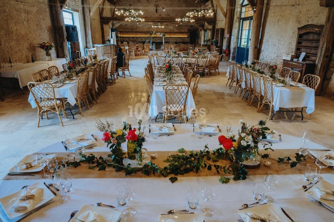 Salle de réception décorée avec des tables dressées, nappes blanches, chaises en rotin et guirlandes végétales.