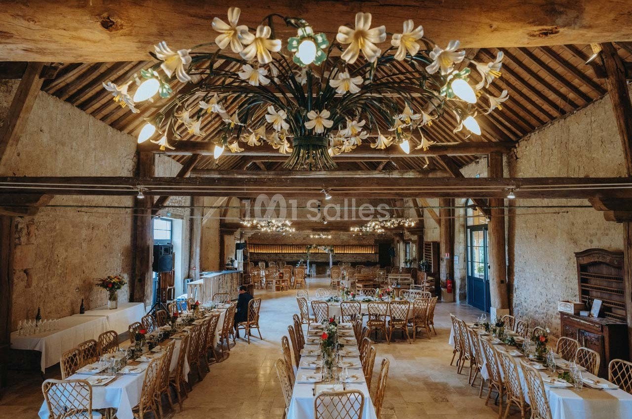 Salle de réception rustique avec tables dressées, chaises en bois et lustres ornés de fleurs blanches.
