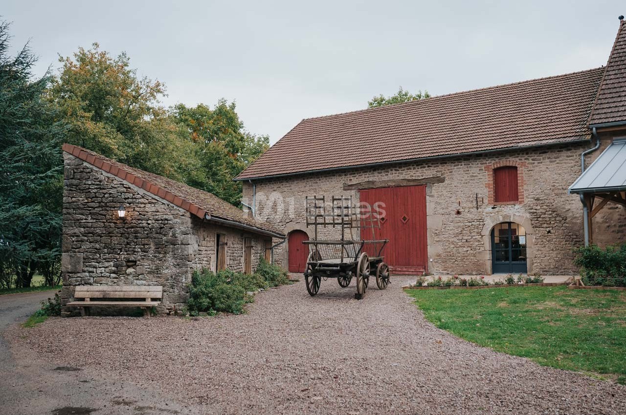 Cour d'une ferme avec une grange en pierre, une charrette en bois et un bâtiment annexe sous un ciel couvert.