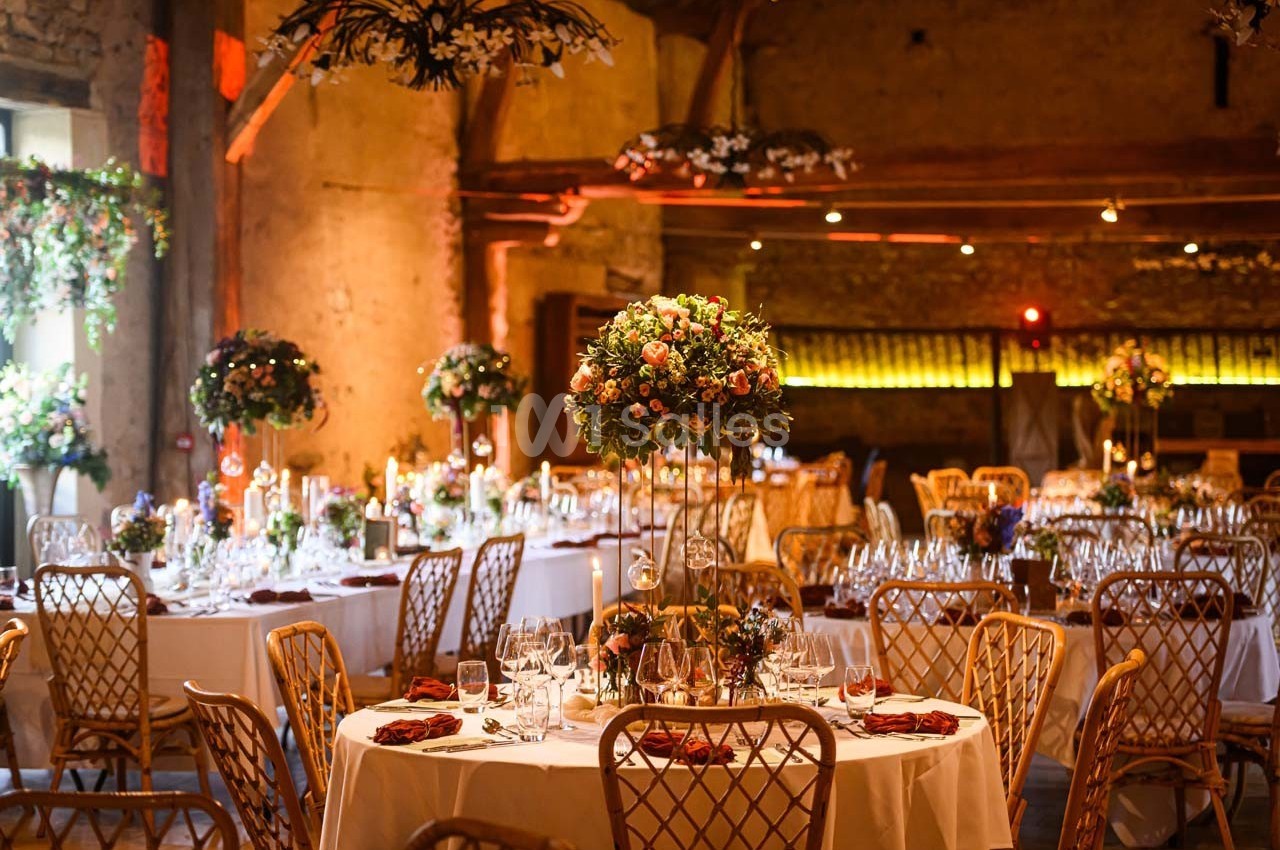 Salle de réception décorée avec des tables rondes, des chaises en bois et des centres de table fleuris sous un éclairage…