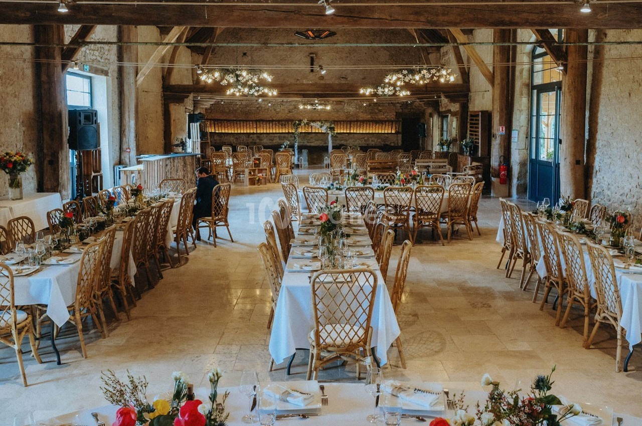 Salle de réception avec tables décorées de nappes blanches, fleurs et chaises en rotin, éclairée par des lustres.