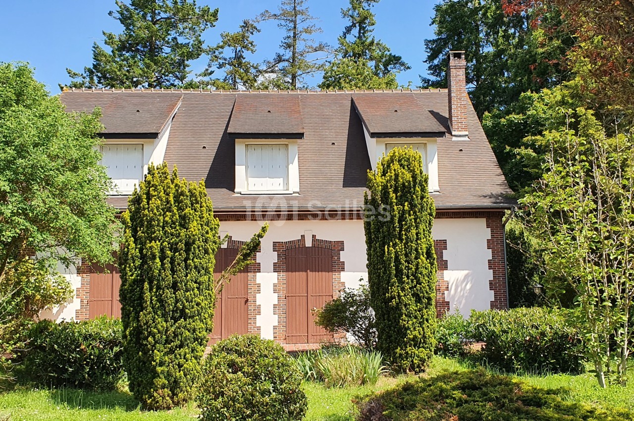 Maison traditionnelle avec toit en pente, volets marron et jardin arboré sous un ciel bleu.