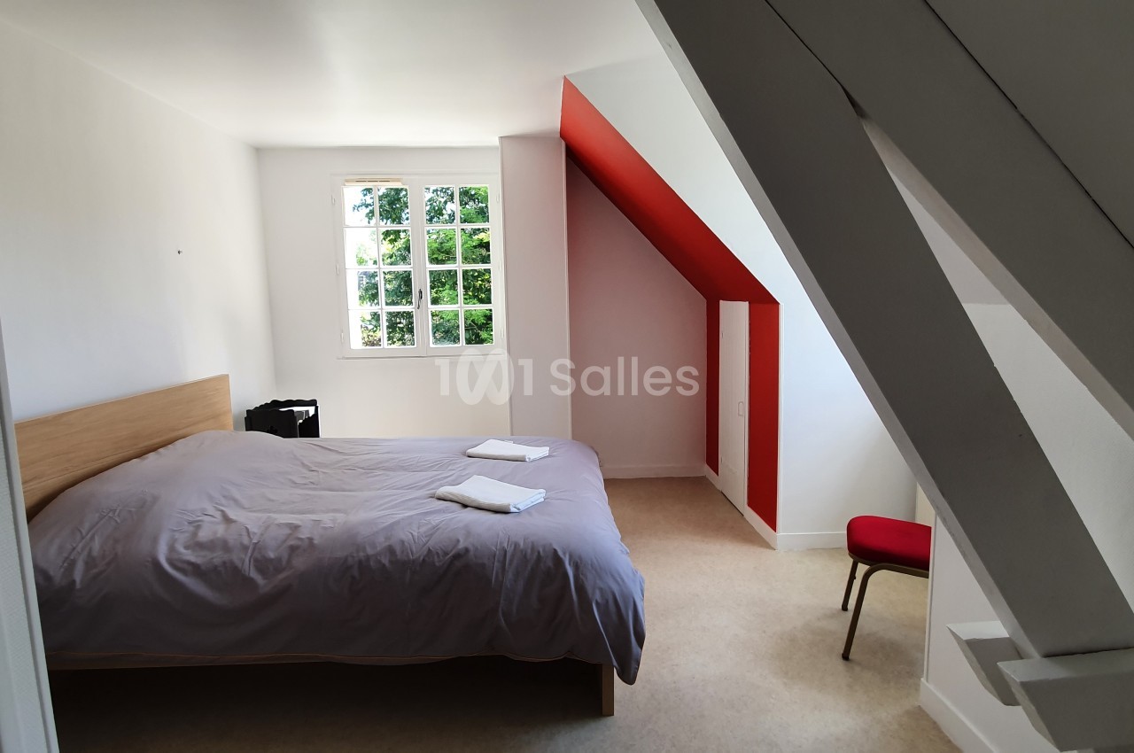 Chambre mansardée lumineuse avec un lit double, draps gris, chaise rouge et fenêtre donnant sur un espace verdoyant.