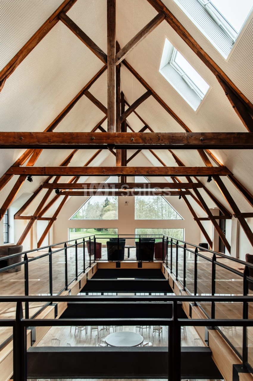 Intérieur d'un bâtiment avec une charpente en bois apparente, mezzanine et grandes fenêtres laissant entrer la lumière…