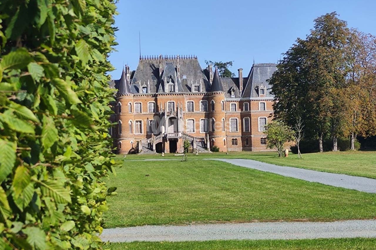 Château en briques rouges avec toits en ardoise, entouré de pelouses et d'arbres, vu depuis une haie.