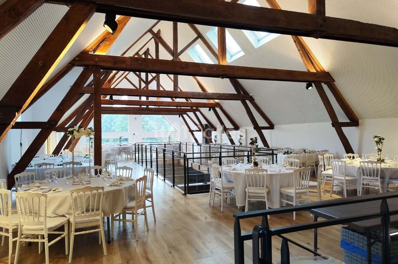 Salle lumineuse avec poutres apparentes, tables rondes dressées pour un événement, parquet en bois clair et baies vitrées.