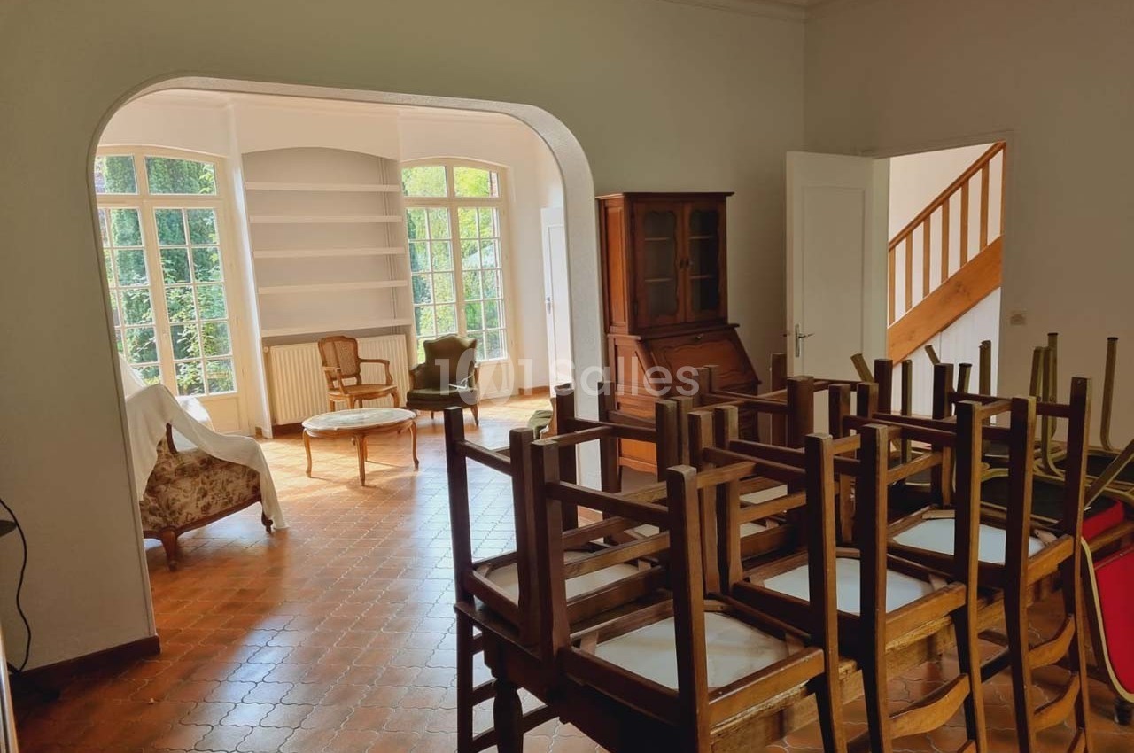 Salle lumineuse avec chaises empilées, sol en terre cuite, meubles en bois et grandes fenêtres donnant sur un jardin.