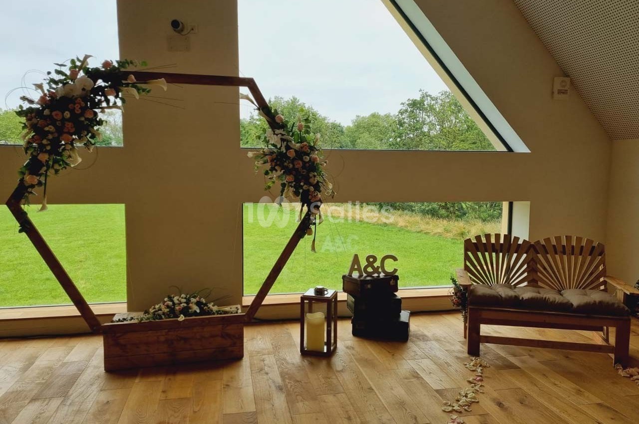 Décoration de mariage avec arche hexagonale fleurie, banc en bois, lanterne et vue sur un jardin à travers une grande…