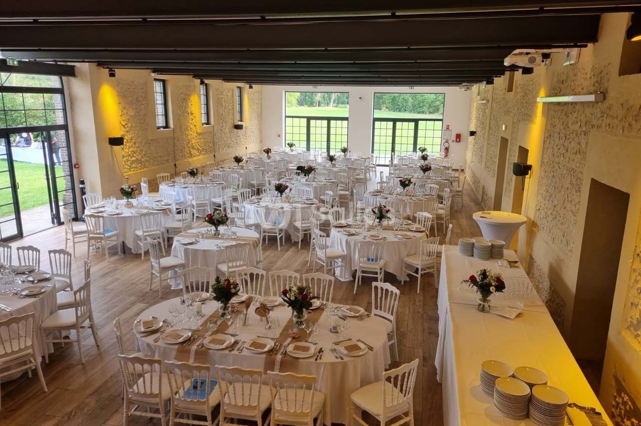 Salle de réception lumineuse avec tables rondes dressées, chaises blanches et décorations florales, vue sur un jardin.