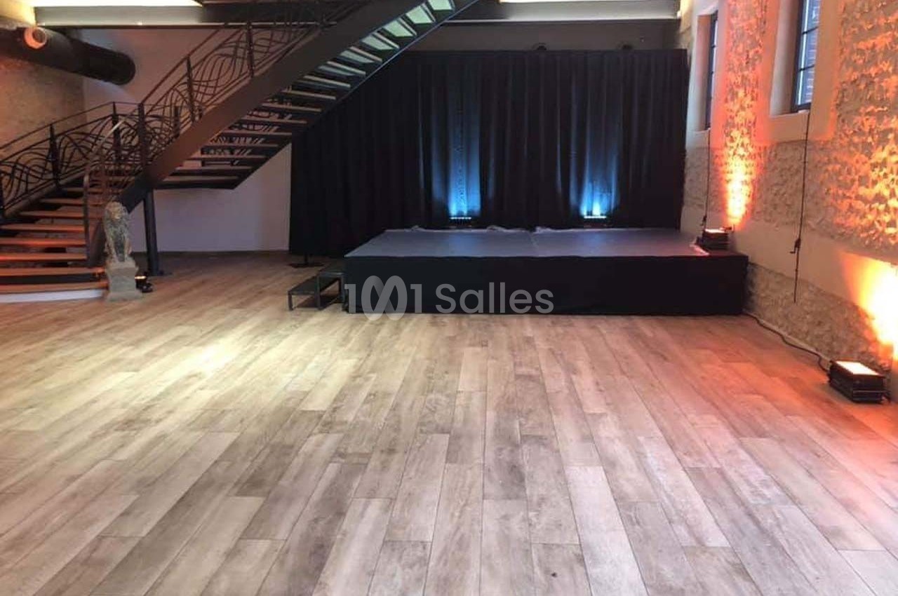 Salle avec parquet clair, petite scène noire, escalier métallique et murs en pierre éclairés par des lumières colorées.