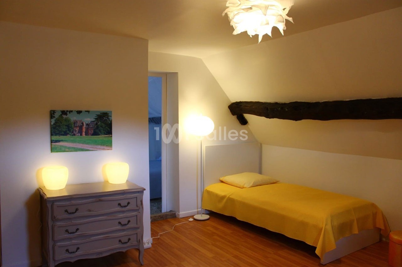 Chambre mansardée avec un lit simple, une commode, deux lampes, un tableau et une poutre apparente.
