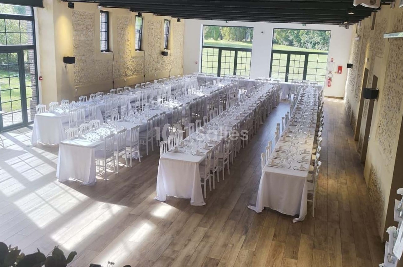 Salle de réception lumineuse avec de longues tables dressées pour un événement, entourée de murs en pierre et grandes…