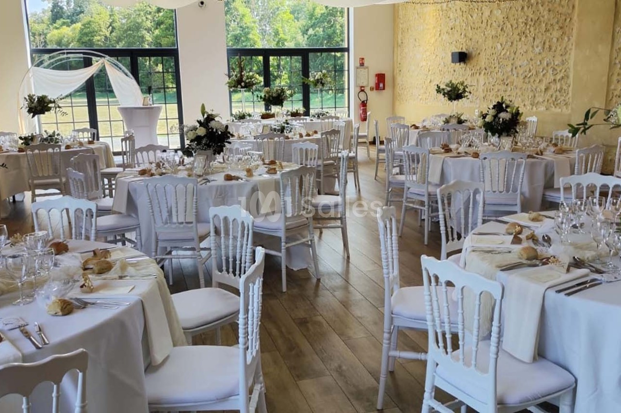 Salle de réception décorée pour un mariage, avec tables rondes dressées, nappes blanches et arrangements floraux.