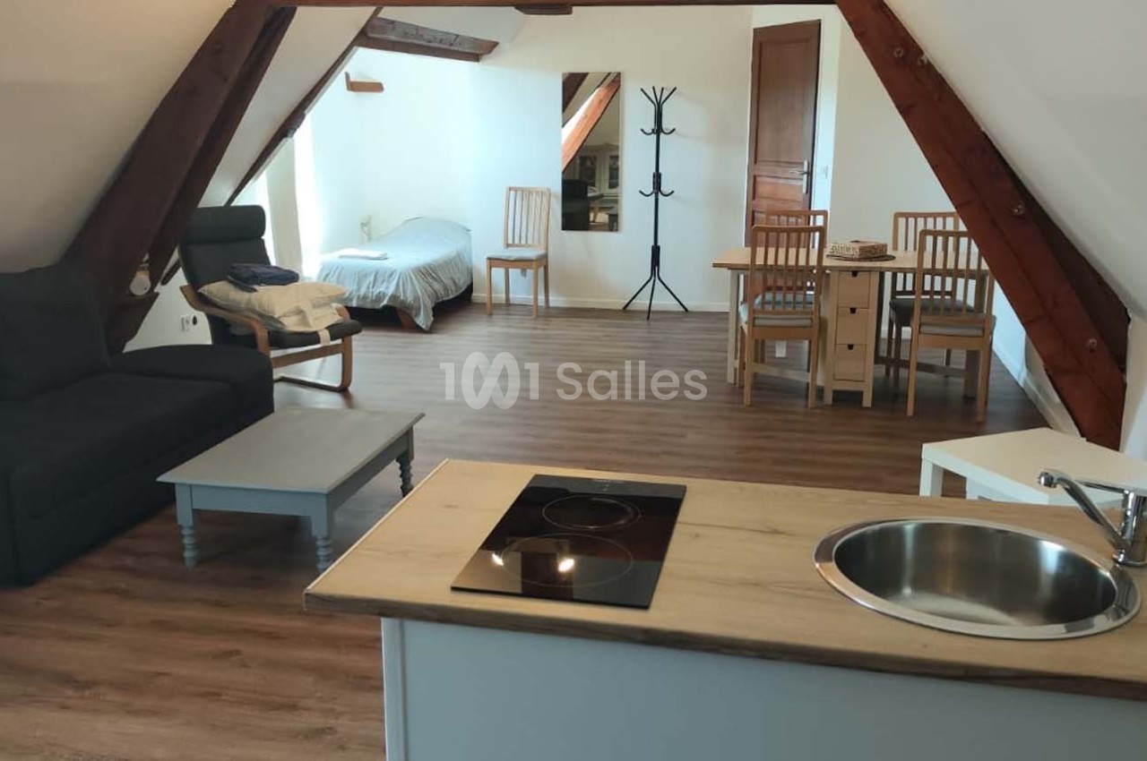 Vue d'un studio mansardé avec coin cuisine, table à manger, canapé, lit simple et sol en parquet.