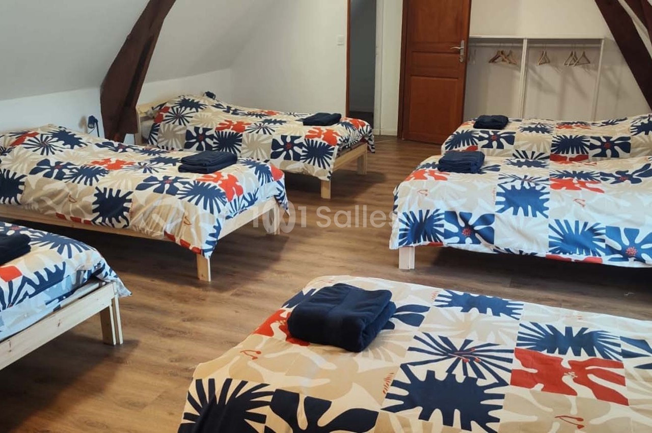 Chambre mansardée avec six lits simples alignés, parure colorée et porte en bois au fond.