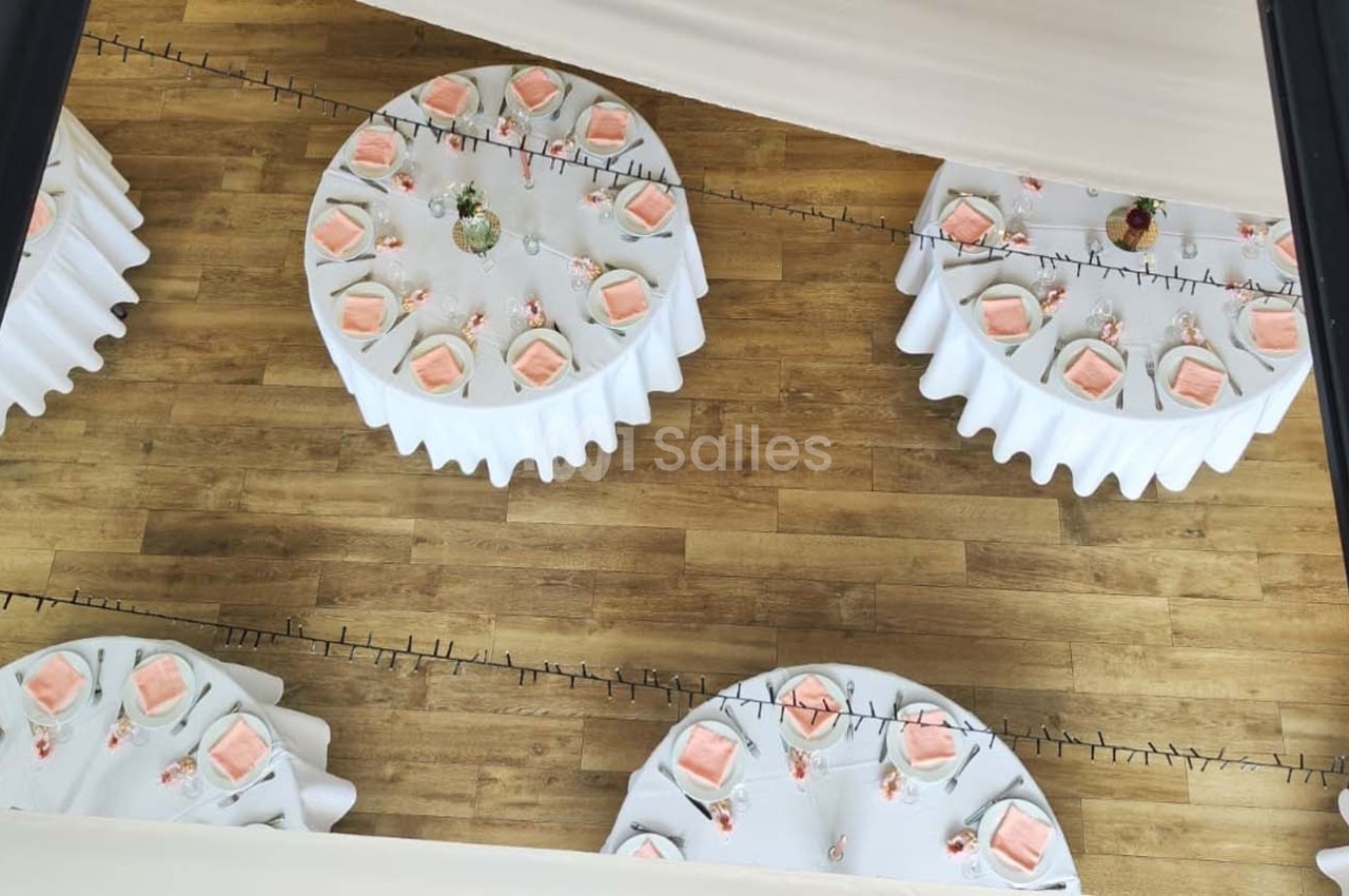 Tables rondes dressées avec nappes blanches, serviettes roses et décorations florales, vues d'en haut sur un sol en bois.
