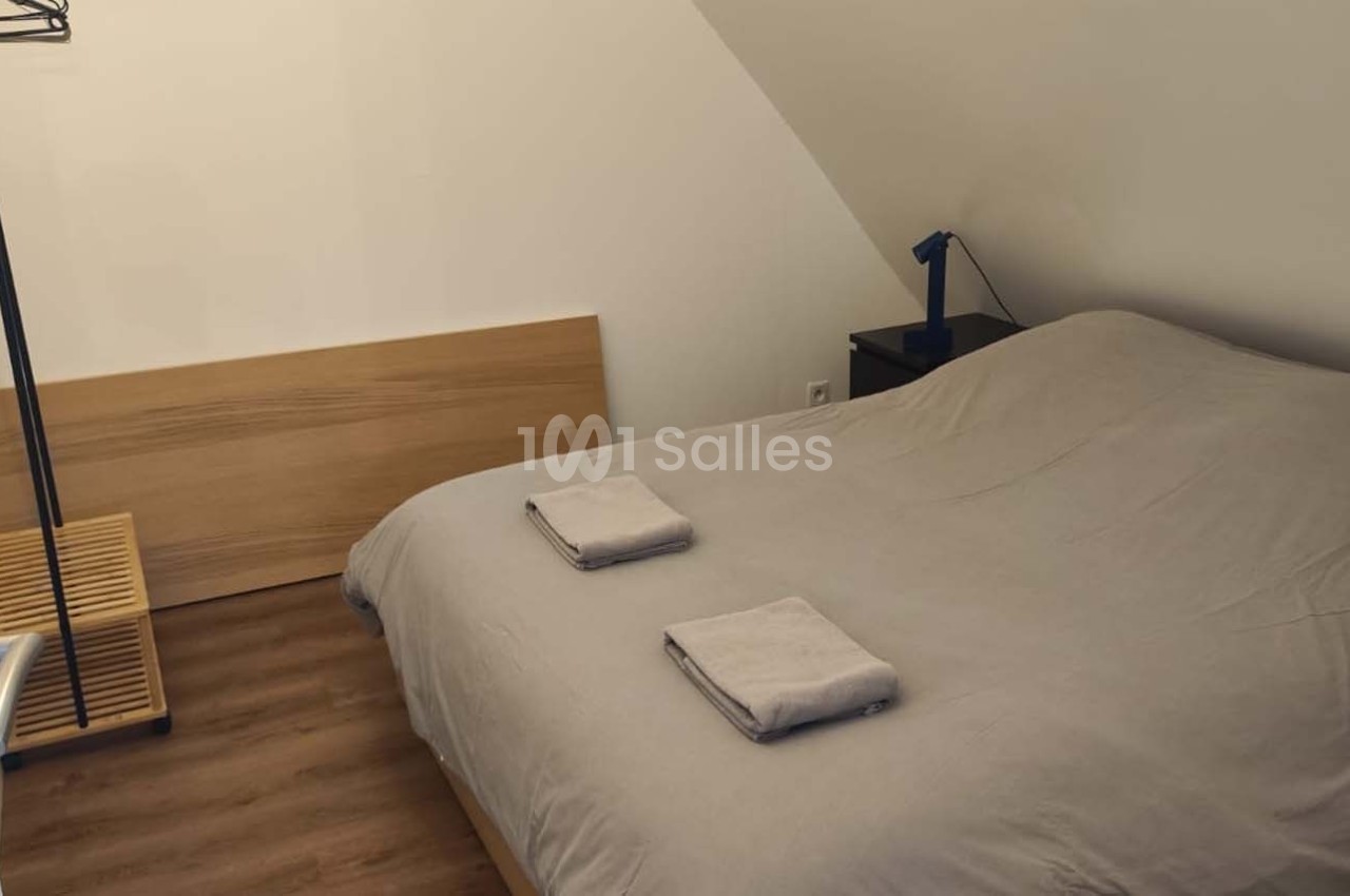 Chambre mansardée avec lit double, draps gris, deux serviettes pliées, lampe de chevet et porte-manteau en métal.