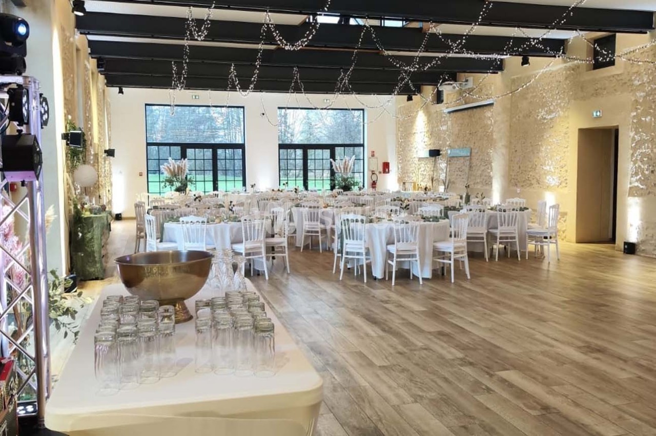 Salle de réception lumineuse avec tables dressées, chaises blanches et décorations suspendues, vue sur un extérieur…