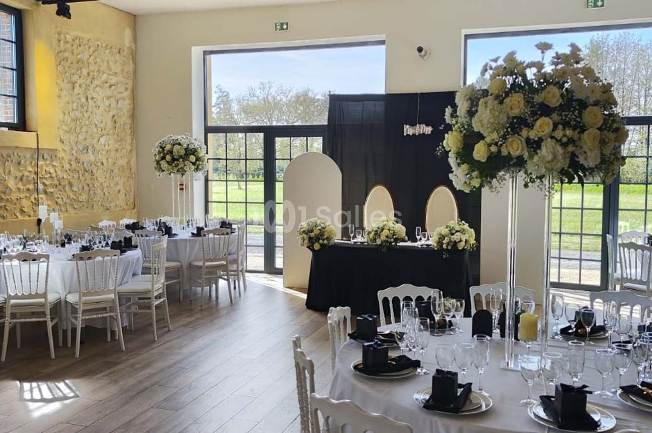 Salle de réception décorée avec des tables dressées, des fleurs blanches et jaunes, et de grandes baies vitrées donnant sur…