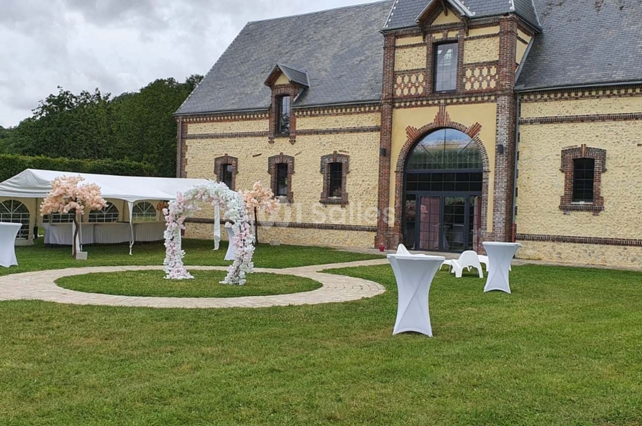 Cour extérieure avec pelouse, arche fleurie, tables hautes et tente blanche devant un bâtiment en briques et pierre.
