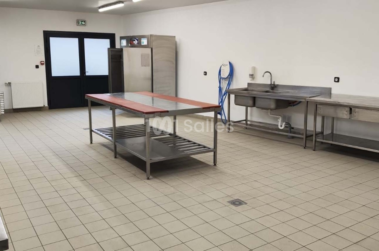 Cuisine professionnelle vide avec équipements en inox, plan de travail central, évier et carrelage beige au sol.