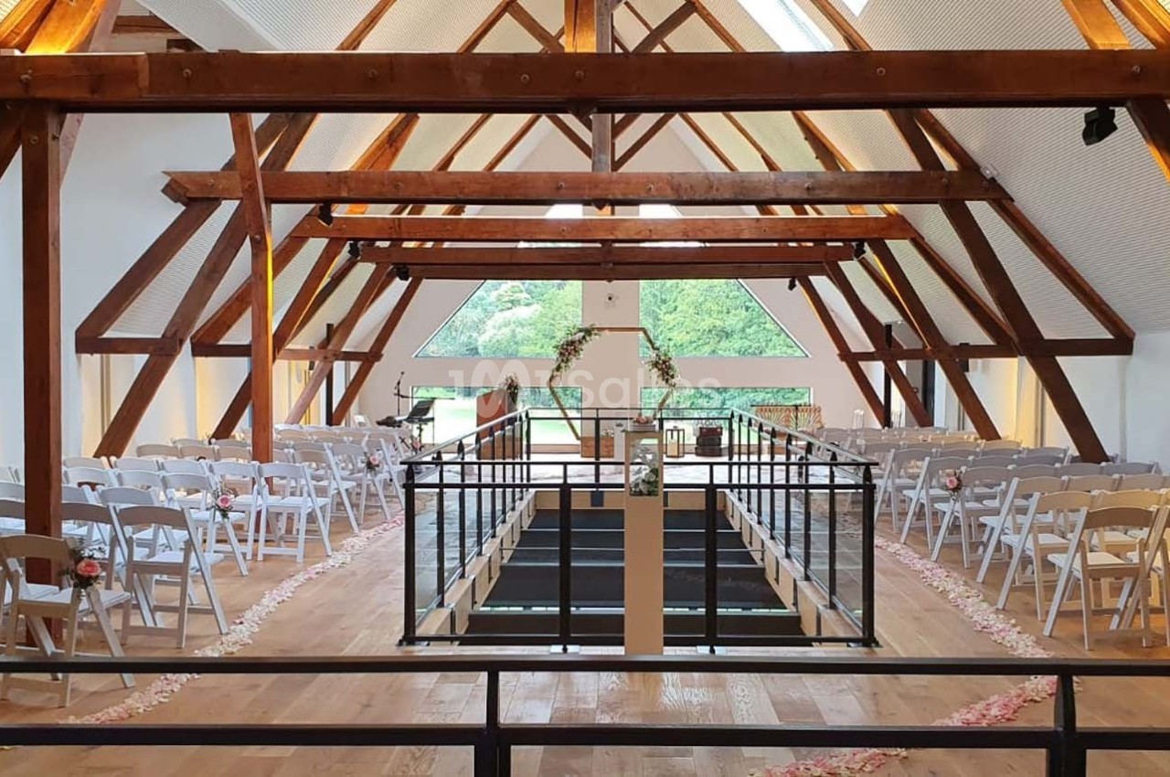 Salle de réception lumineuse avec charpente en bois, rangées de chaises blanches et allée centrale décorée de pétales.