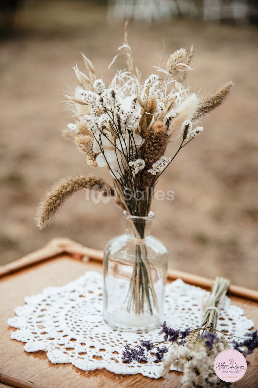 Bouquet de fleurs séchées dans un vase en verre, posé sur une nappe en dentelle sur une table en bois.