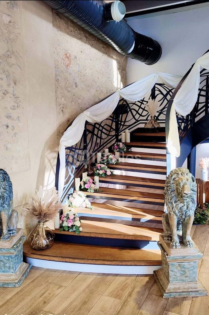 Escalier en bois décoré de fleurs et drapés blancs, encadré par deux statues de lions dans une salle au mur en pierre.
