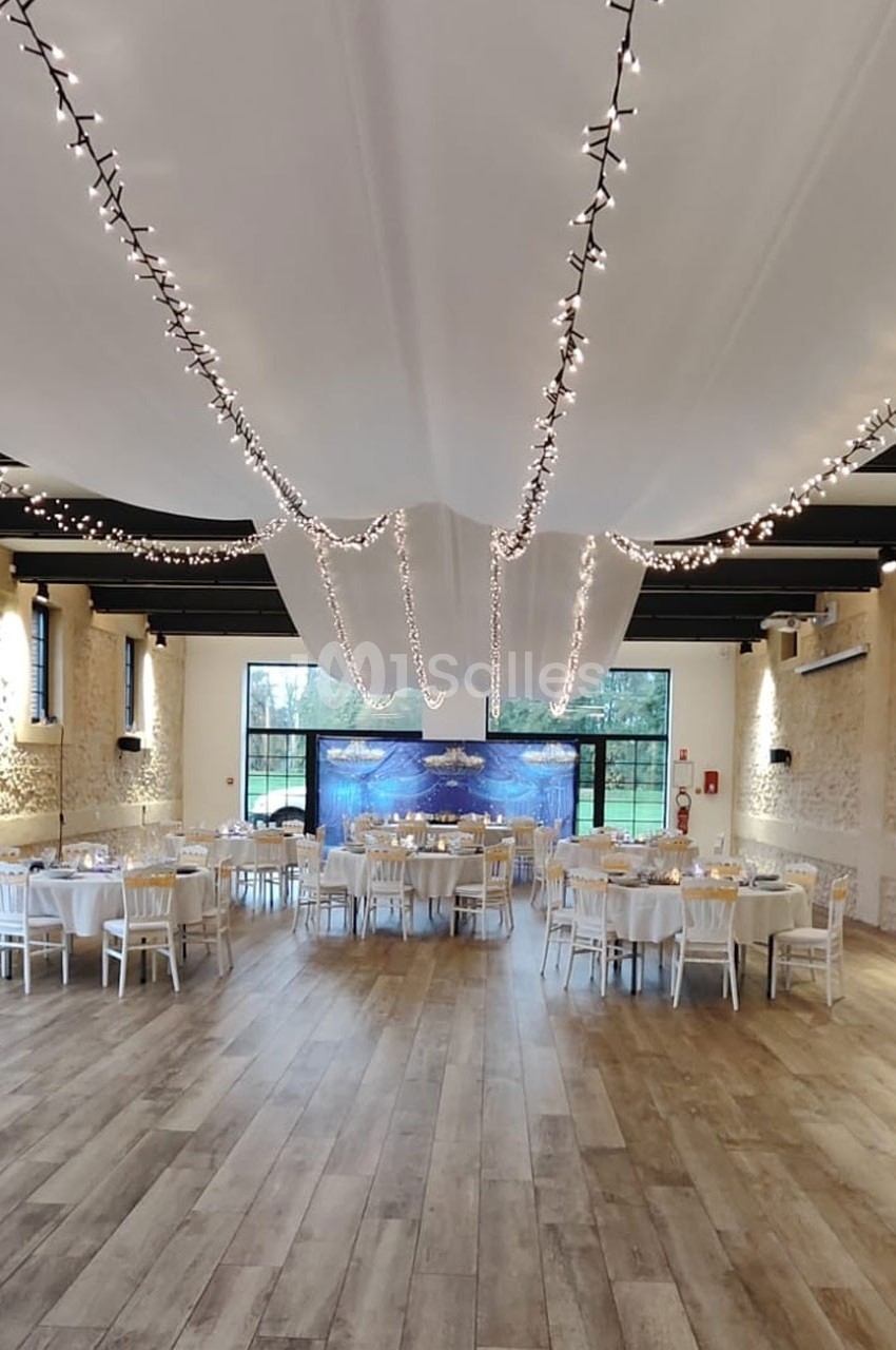 Salle de réception lumineuse avec tables dressées, guirlandes lumineuses suspendues et vue sur un jardin à travers de…