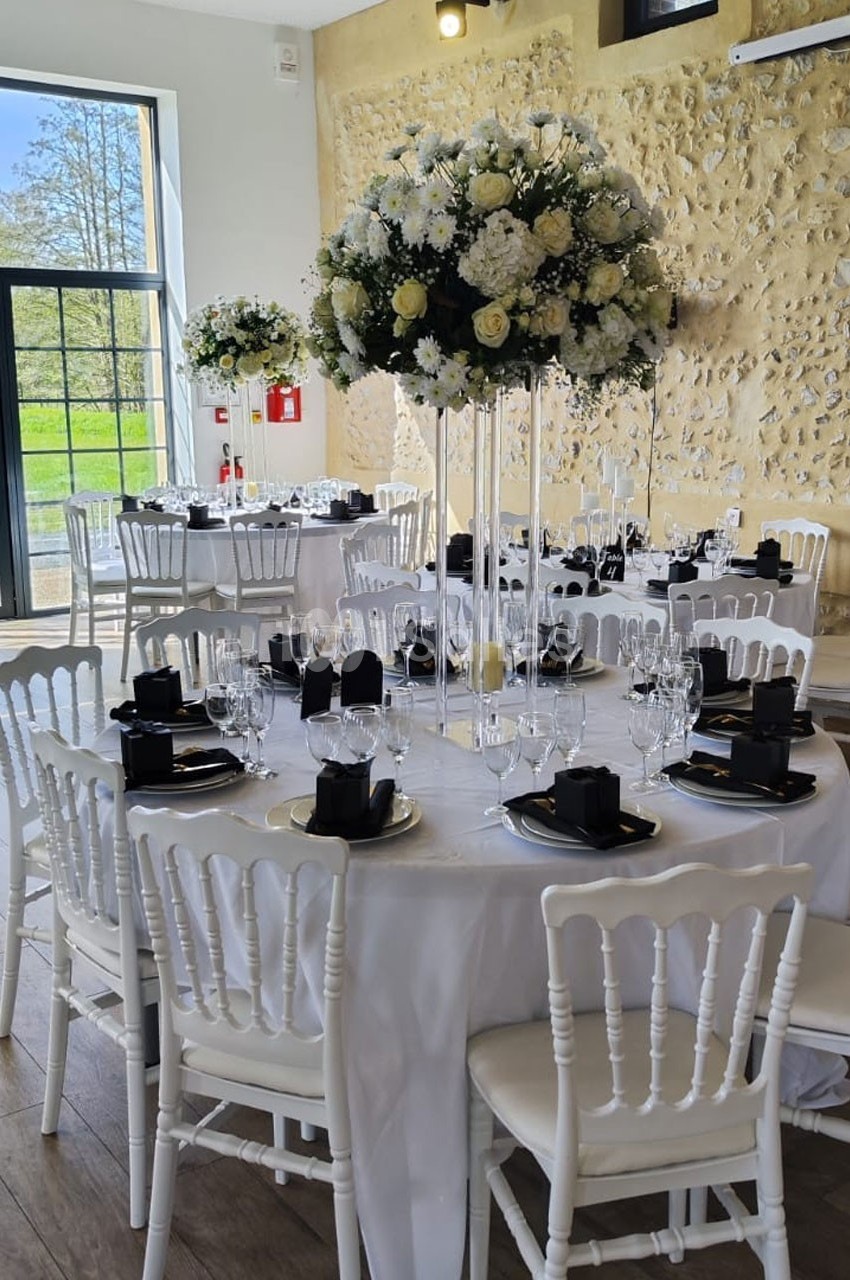 Salle de réception décorée avec des tables rondes, nappes blanches, chaises blanches et centres de table fleuris.