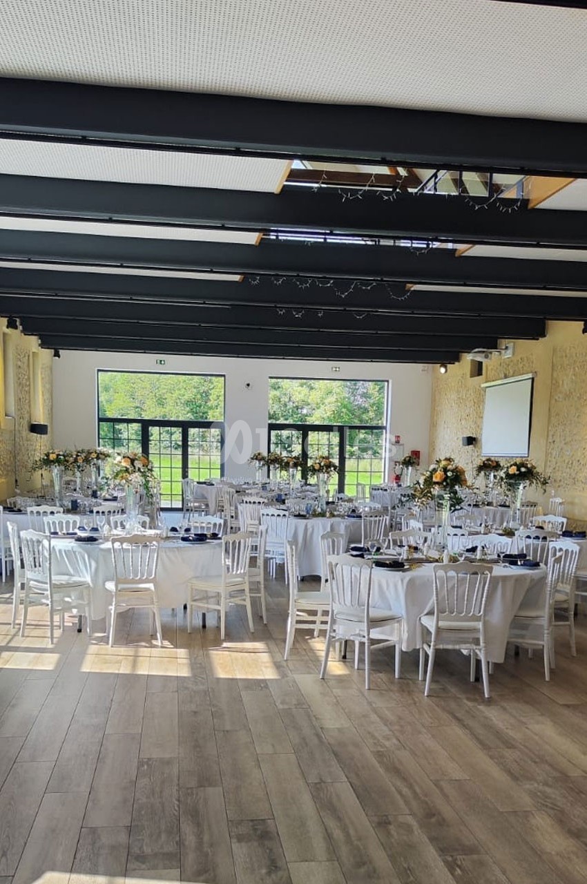 Salle de réception lumineuse avec tables décorées, chaises blanches et grandes baies vitrées donnant sur un espace vert.