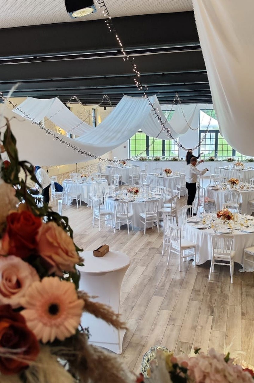 Salle de réception décorée avec des tables rondes, nappes blanches, fleurs et guirlandes lumineuses suspendues.