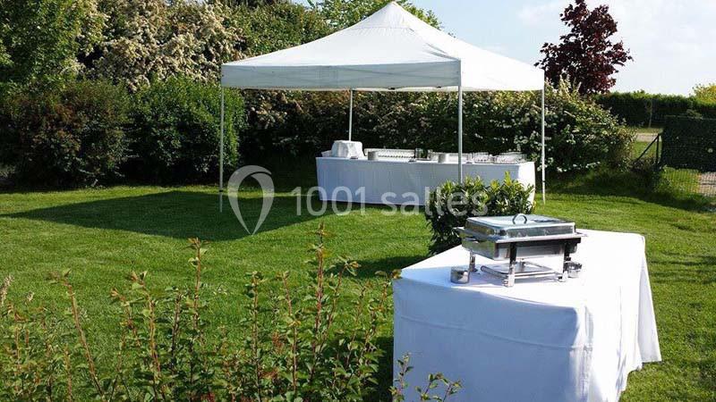 Tente blanche avec tables dressées pour un buffet en extérieur, sur une pelouse entourée de végétation.