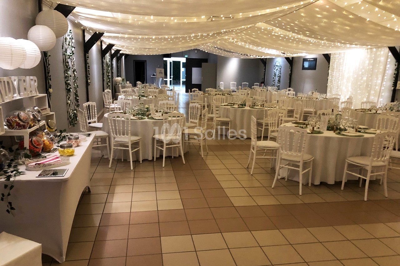 Salle de réception décorée avec des tables rondes, chaises blanches, guirlandes lumineuses et un buffet sur la gauche.