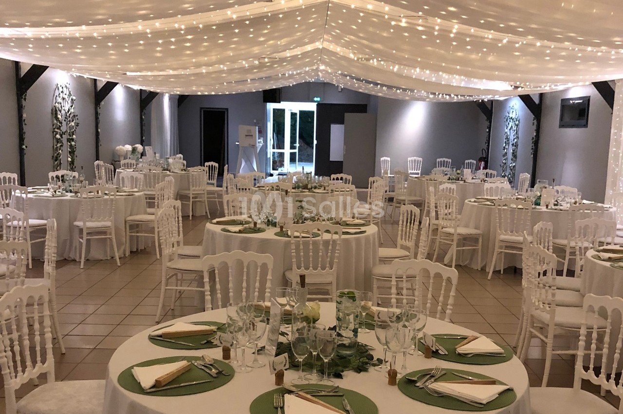 Salle de réception décorée avec des tables rondes, nappes blanches, chemins de table verts et guirlandes lumineuses…