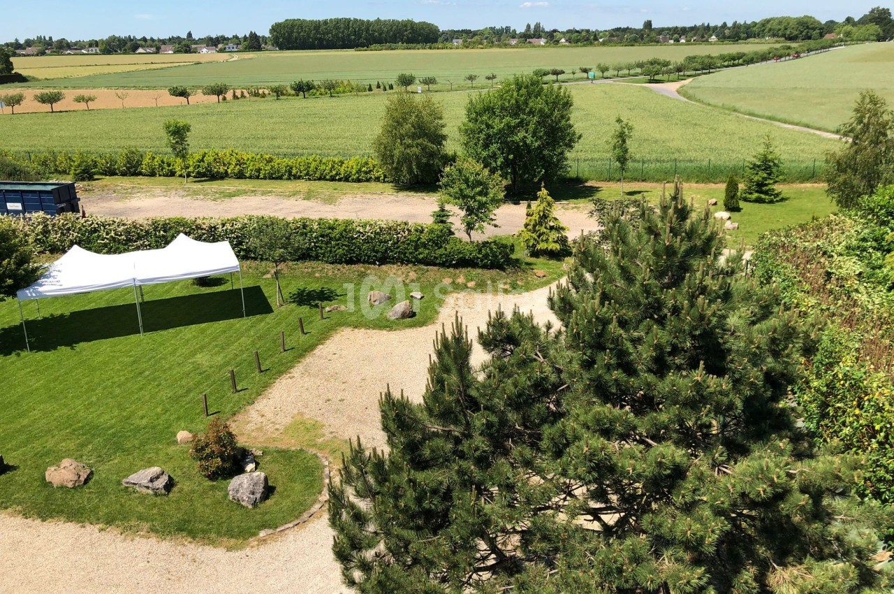 Vue aérienne d'un jardin avec pelouse, arbres, tente blanche et champs verdoyants à l'arrière-plan.