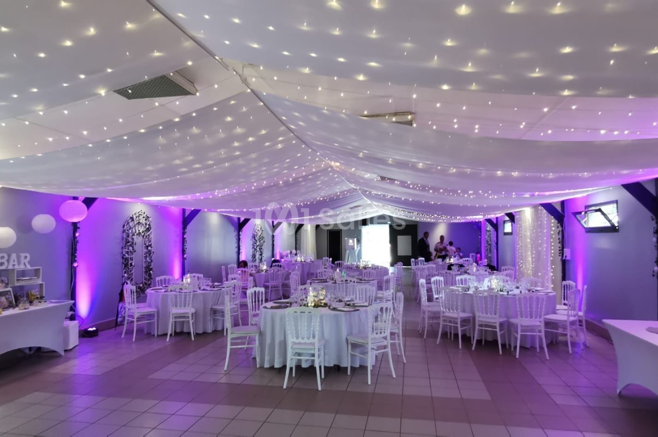 Salle de réception décorée avec des guirlandes lumineuses au plafond, tables rondes dressées et éclairage violet.