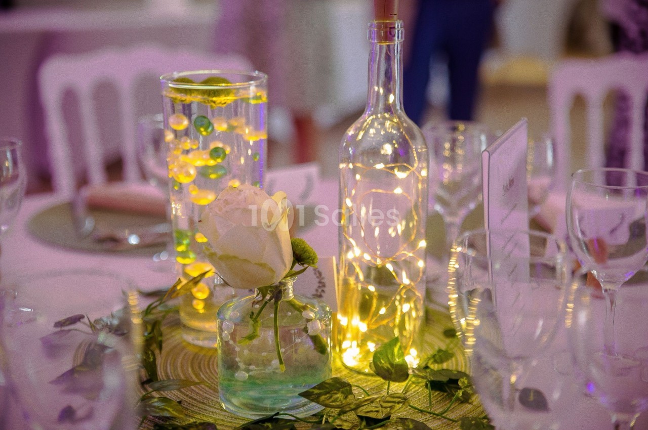Centre de table décoratif avec une rose blanche, des guirlandes lumineuses et des vases en verre sur une nappe verte.