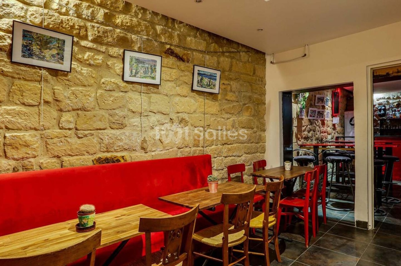 Salle de restaurant avec murs en pierre, banquette rouge, tables en bois et vue sur un bar éclairé en rouge.