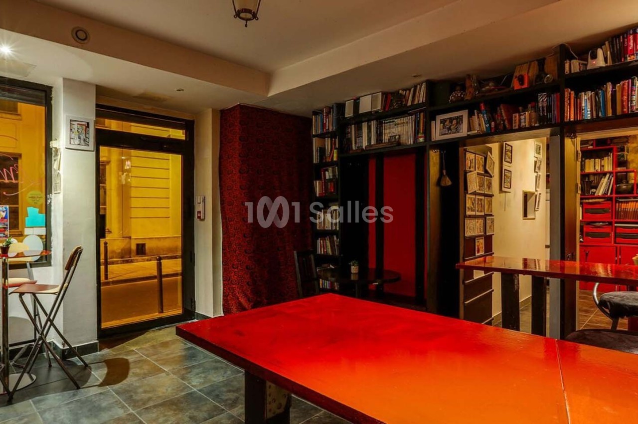 Intérieur d'un café avec tables rouges, chaises hautes, étagères remplies de livres et lumière chaleureuse.