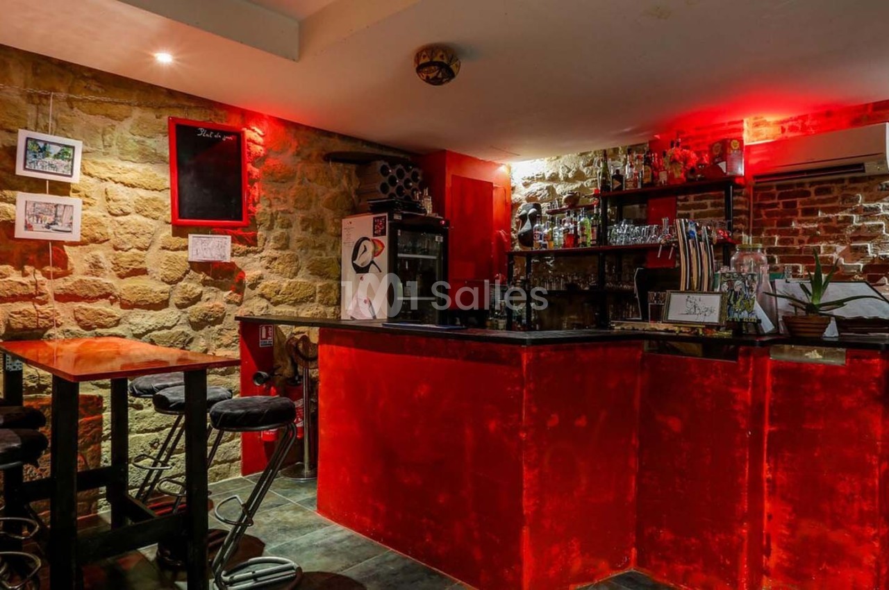 Bar avec comptoir rouge éclairé, tabourets hauts, murs en pierre et briques, décorations murales et étagères avec bouteilles.