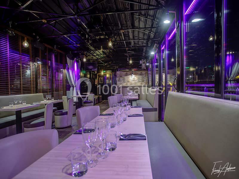 Salle de restaurant moderne avec éclairage violet, tables dressées et banquettes claires le long des baies vitrées.