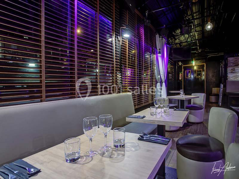 Salle de restaurant moderne avec banquettes claires, tables dressées et éclairage violet tamisé.