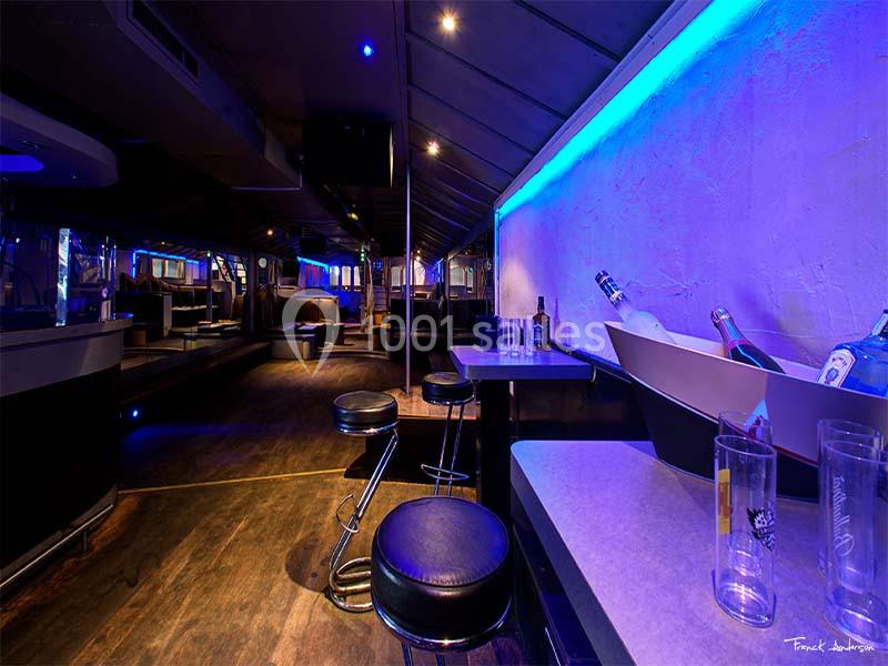 Intérieur d'un bar avec comptoir, tabourets, bouteilles et éclairage bleu dans une ambiance nocturne.