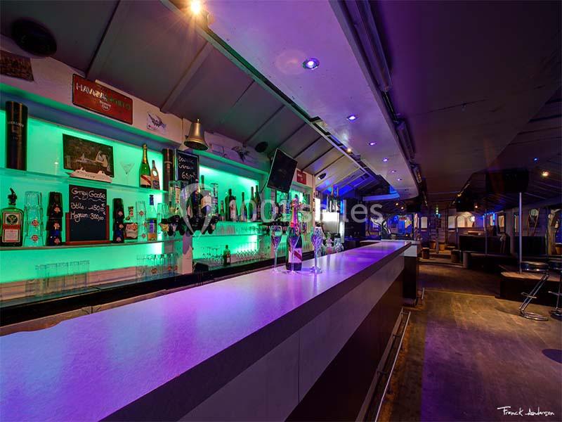 Bar moderne avec éclairage coloré, comptoir long et rangées de bouteilles alignées sur des étagères rétroéclairées.