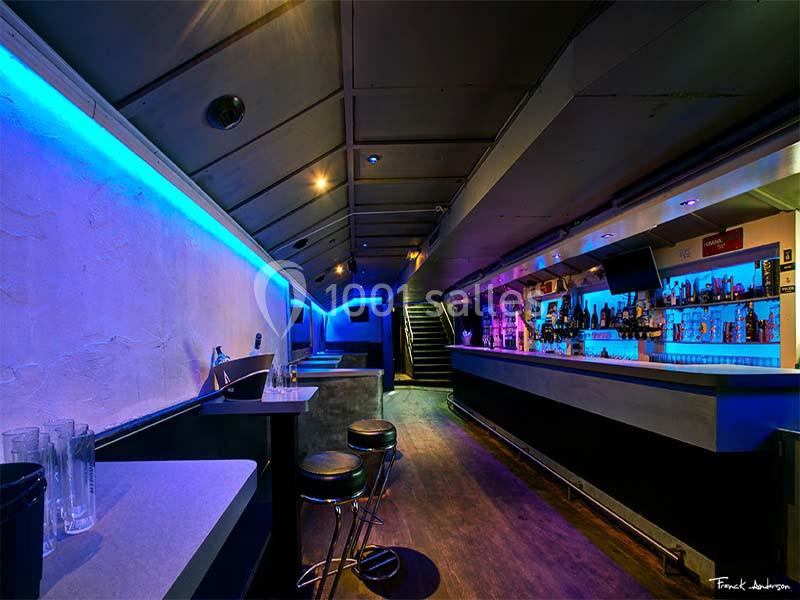 Intérieur d'un bar avec un éclairage bleu, un comptoir à droite et des tables hautes avec tabourets à gauche.